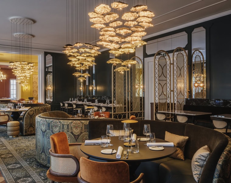 Elegantes Restaurant mit dunkelgrünen Wänden, opulenten goldenen Blüten-Kronleuchtern, gemusterten Polstersofas, samtbezogenen Sesseln und gedeckten Tischen auf orientalischem Teppich.