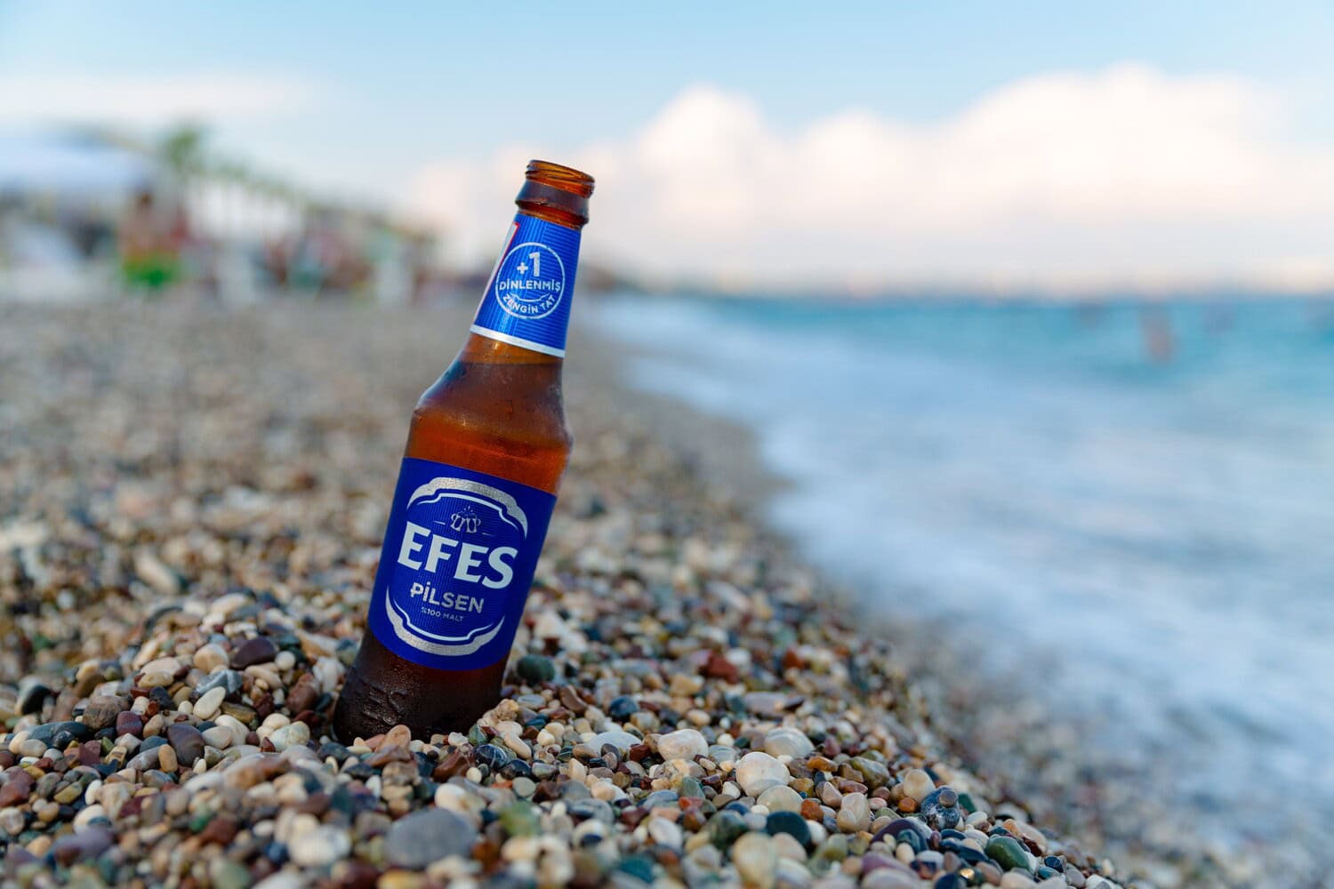 Flasche Efes-Pilsen-Bier steht im Kiesstrand, im Hintergrund das Meer.