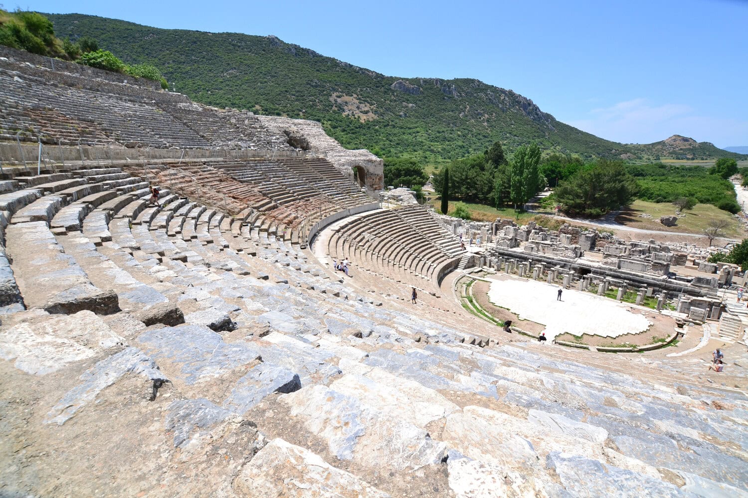 Grosses antikes Theater von Ephesus mit ansteigenden Steinraengen vor bewaldeten Huegeln.