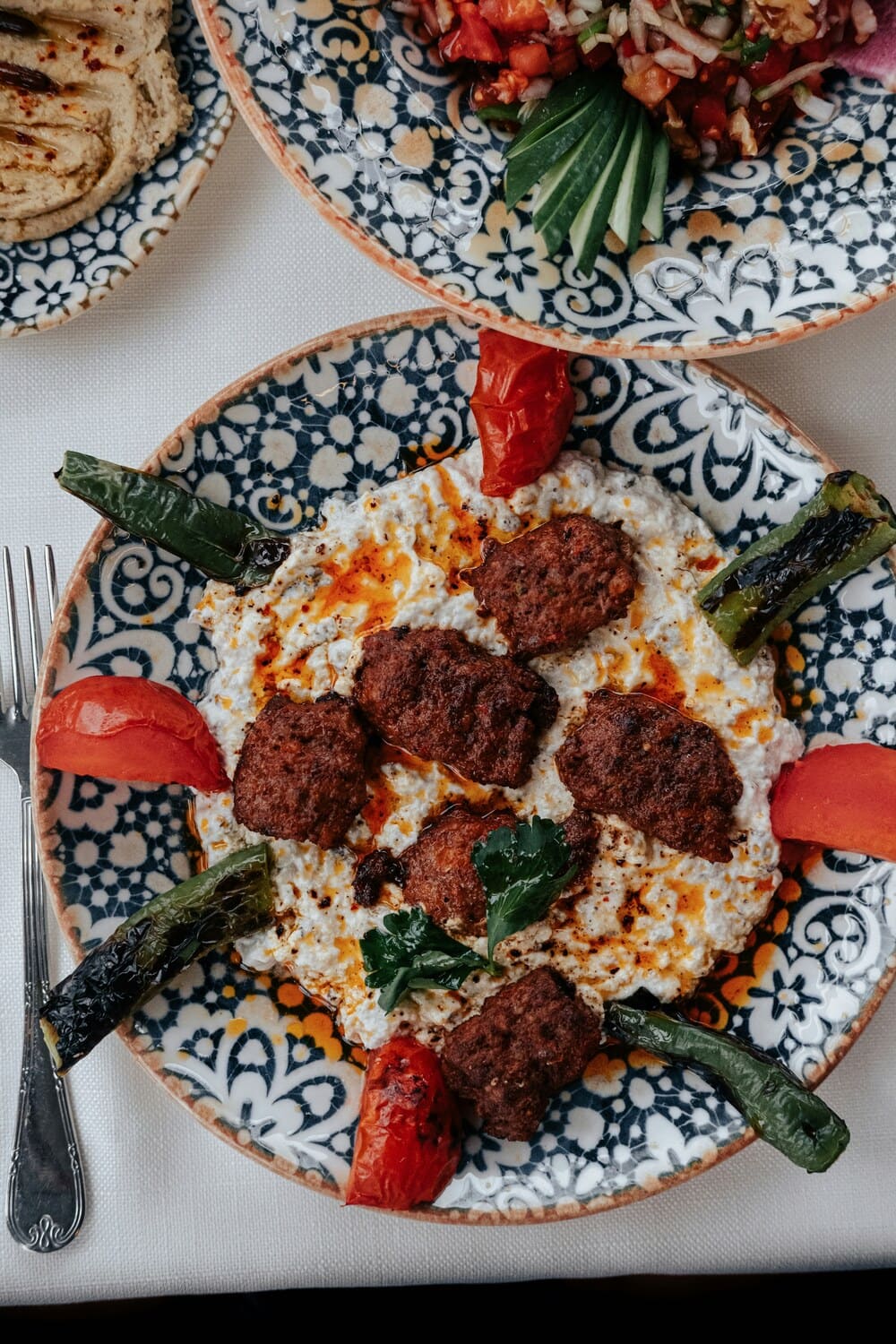 Teller mit gegrillten Koefte auf Joghurt, dazu geroestete Tomaten und Paprika, im Hintergrund Mezze.