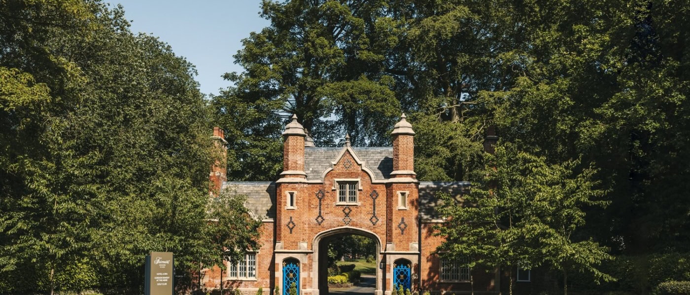 Historisches Torhaus aus rotem Backstein im Tudorstil mit spitzem Mittelbogen, eingerahmt von hohen grünen Bäumen, am Ende einer Zufahrtsstraße bei Sonnenschein.