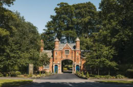 Historisches Torhaus aus rotem Backstein im Tudorstil mit spitzem Mittelbogen, eingerahmt von hohen grünen Bäumen, am Ende einer Zufahrtsstraße bei Sonnenschein.