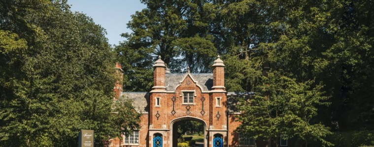Historisches Torhaus aus rotem Backstein im Tudorstil mit spitzem Mittelbogen, eingerahmt von hohen grünen Bäumen, am Ende einer Zufahrtsstraße bei Sonnenschein.