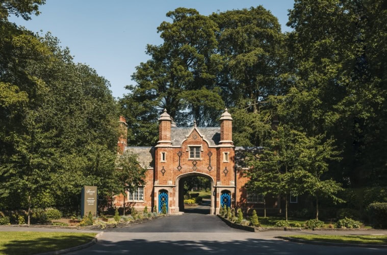 Historisches Torhaus aus rotem Backstein im Tudorstil mit spitzem Mittelbogen, eingerahmt von hohen grünen Bäumen, am Ende einer Zufahrtsstraße bei Sonnenschein.