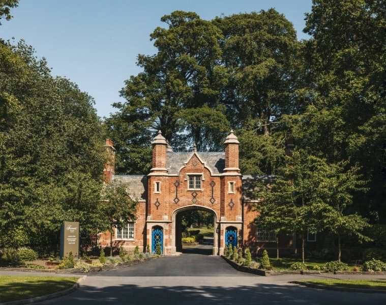 Historisches Torhaus aus rotem Backstein im Tudorstil mit spitzem Mittelbogen, eingerahmt von hohen grünen Bäumen, am Ende einer Zufahrtsstraße bei Sonnenschein.