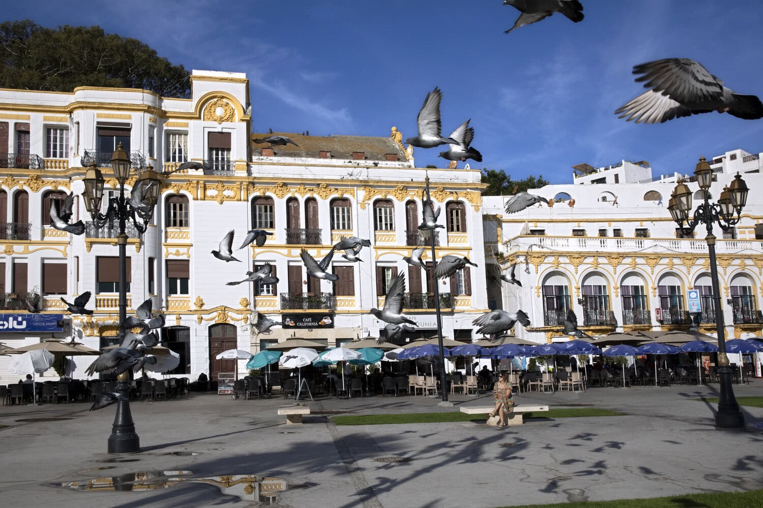 Auffliegende Tauben auf einem Platz in Tanger vor weiss-goldenen Altstadtgebaeuden mit Cafe-Terrassen.