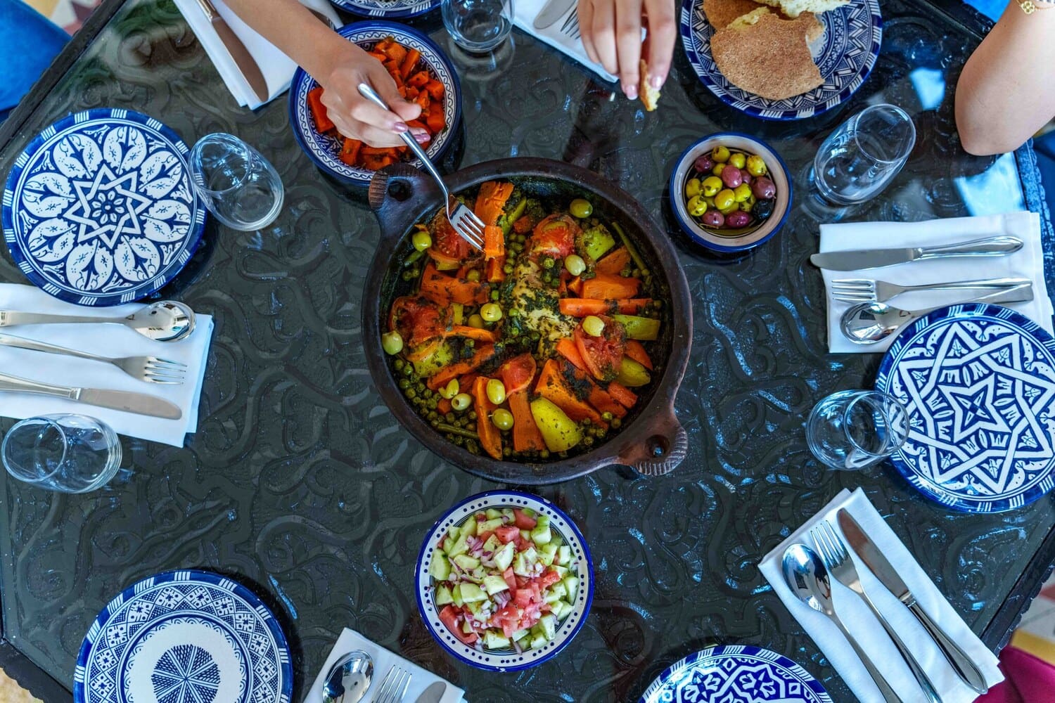 Marokkanisches Tajine-Gericht mit Gemuese in der Tischmitte, umgeben von blau gemustertem Geschirr.