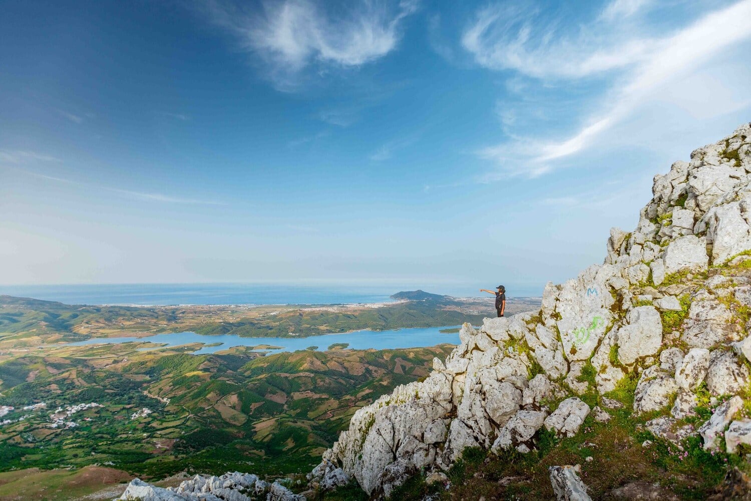 Wanderer auf einem Felsgrat mit weitem Blick ueber gruene Landschaft, See und Meer bei Tanger.