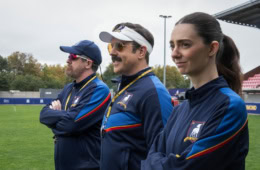 Szene aus Ted Lasso: Drei Trainer in blauen Trainingsjacken stehen nebeneinander am Spielfeldrand.
