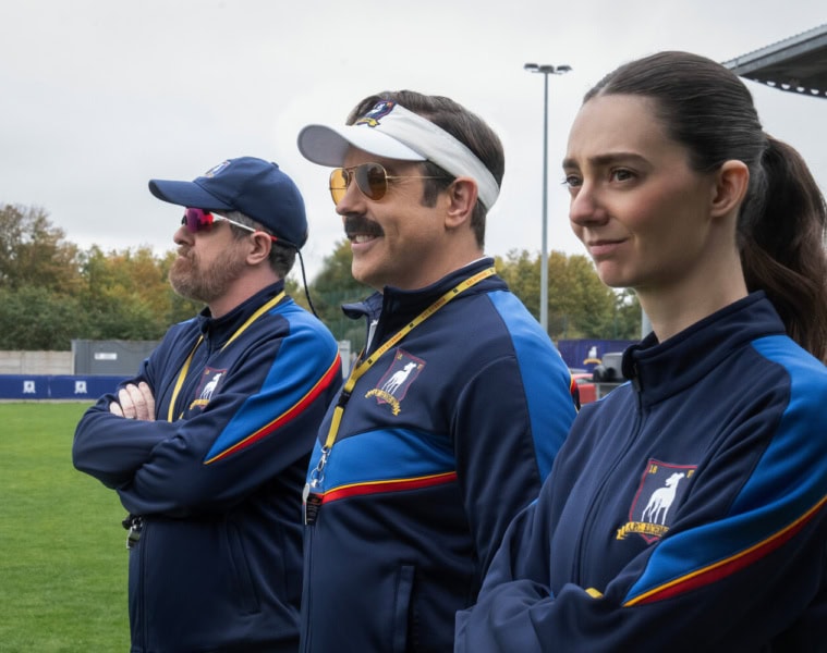 Szene aus Ted Lasso: Drei Trainer in blauen Trainingsjacken stehen nebeneinander am Spielfeldrand.