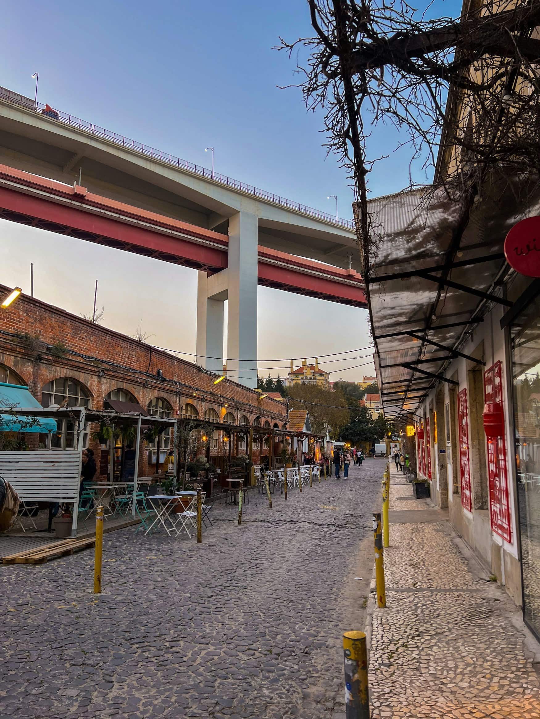 Gasse mit Cafés und Lagerhallen unter der Ponte 25 de Abril im Lissabonner Viertel Alcântara