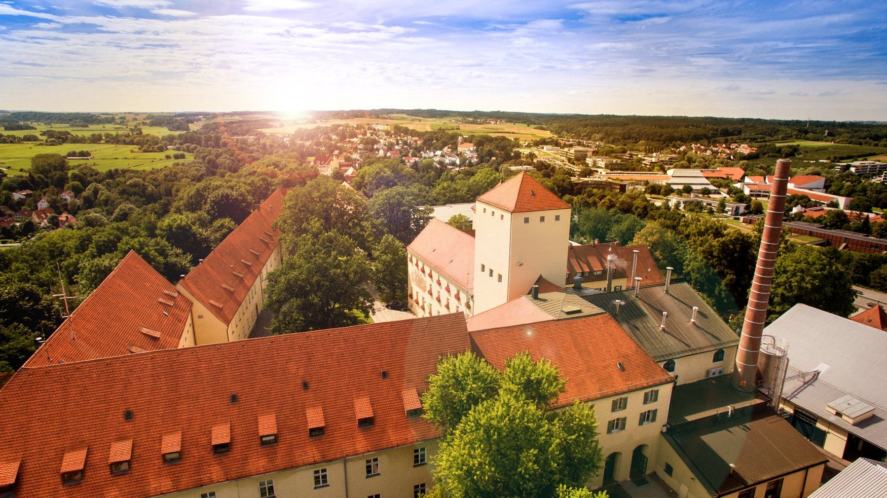 Luftaufnahme der Brauerei Weihenstephan mit ihren historischen Gebäuden mit roten Dächern und dem markanten Schornstein, umgeben von Bäumen und Feldern im Gegenlicht der untergehenden Sonne.