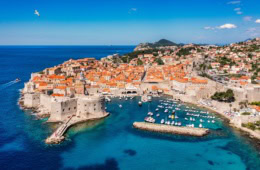 Luftaufnahme von Dubrovnik an einem sonnigen Tag, links die Adria, mittig die Stadt mit Stadtmauer und Hafenbecken