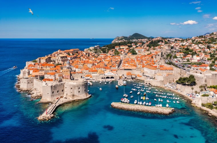 Luftaufnahme von Dubrovnik an einem sonnigen Tag, links die Adria, mittig die Stadt mit Stadtmauer und Hafenbecken