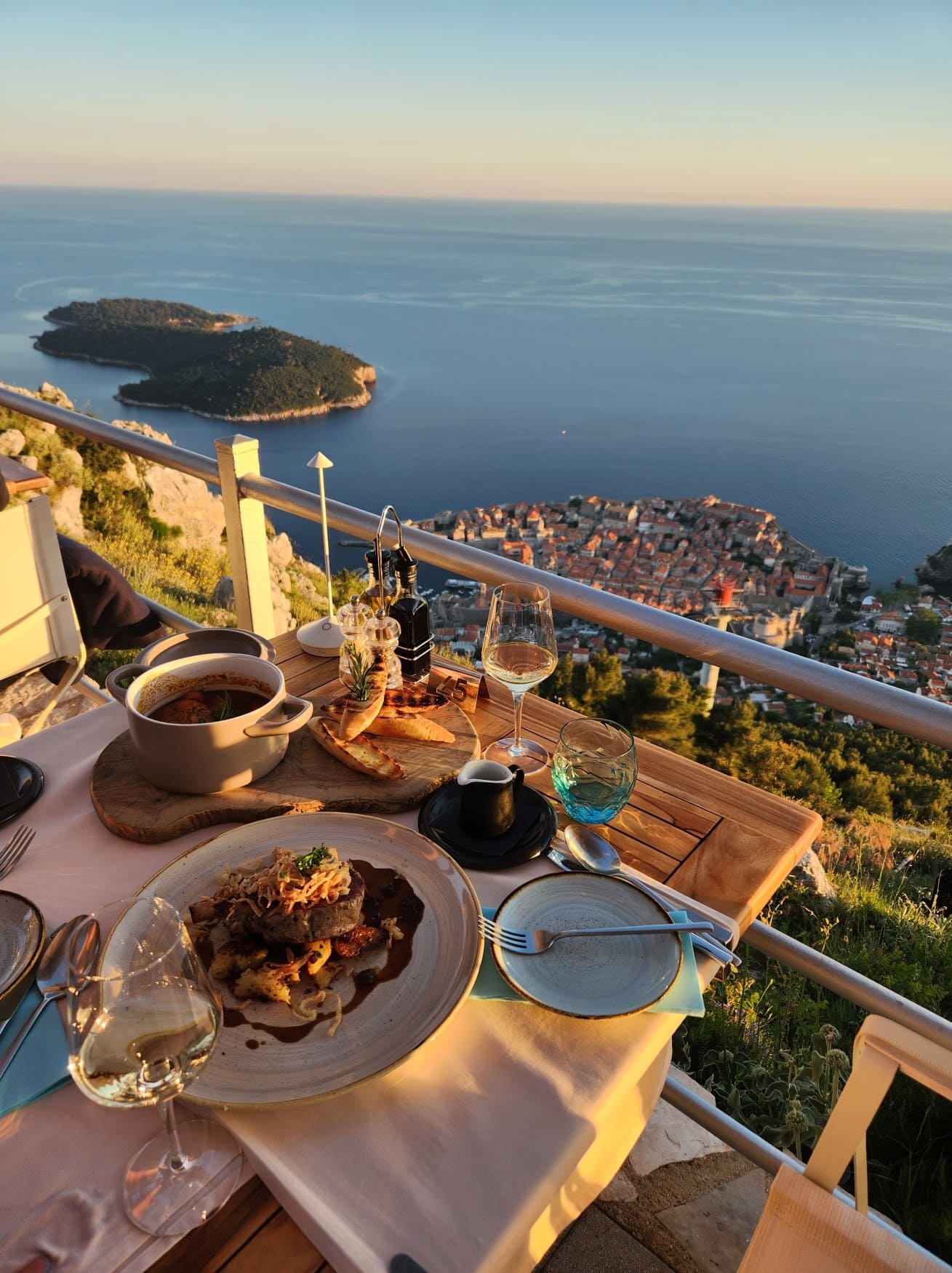 Ein gedeckter Tisch für zwei Personen auf der Terrasse eines eleganten Hotels, direkt an einem Geländer. Dahinter geht der Blick hinab auf Dubrovnik und die Adria im Licht des Sonnenuntergangs.