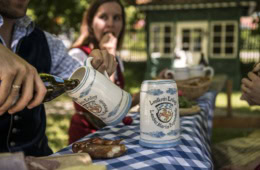 Hand gießt Bier aus einer Flasche in einen bemalten Steinkrug mit dem Wappen des Landkreises Erding, auf dem Tisch liegt eine Brezel, im Hintergrund sitzt eine Frau in Tracht.