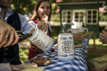 Hand gießt Bier aus einer Flasche in einen bemalten Steinkrug mit dem Wappen des Landkreises Erding, auf dem Tisch liegt eine Brezel, im Hintergrund sitzt eine Frau in Tracht.
