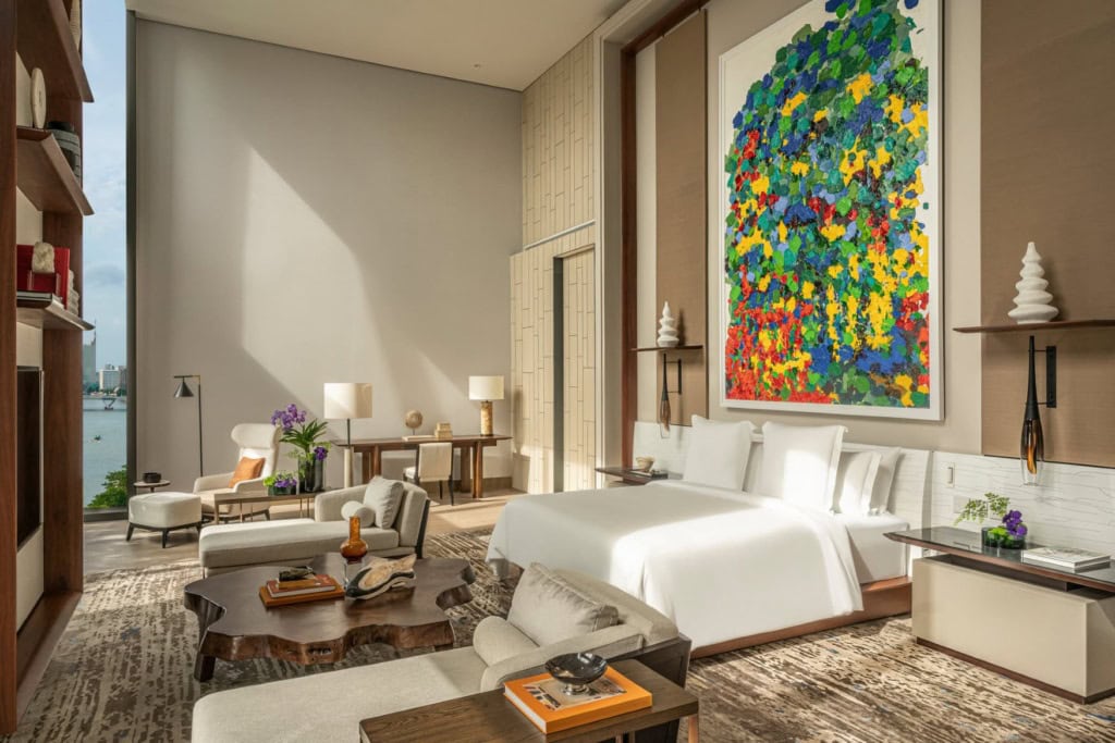 Blick in eine gro&szlig;e, moderne Hotelsuite, mittig ein breites Bett, dar&uuml;ber ein gut zwei Meter hohes, modernes Gem&auml;lde. Vor dem Bett ein aus einem Baumstamm geschnittener Couchtisch, zu den Seiten je eine Recamiere.