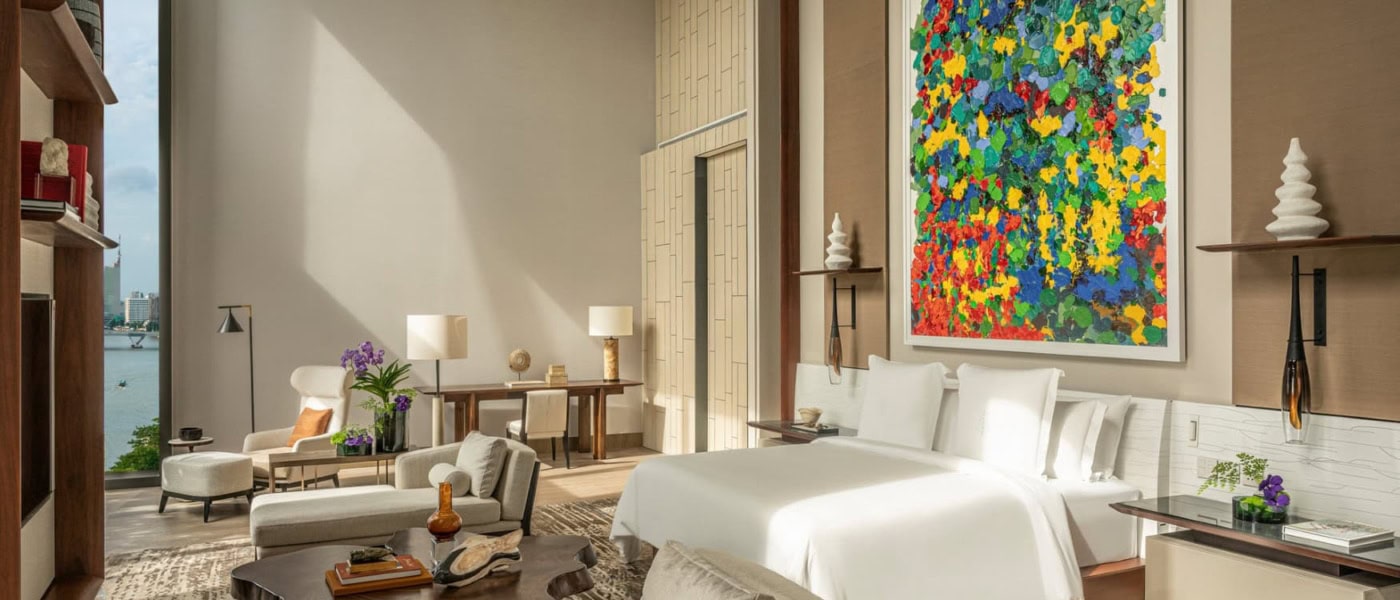 Blick in eine große, moderne Hotelsuite, mittig ein breites Bett, darüber ein gut zwei Meter hohes, modernes Gemälde. Vor dem Bett ein aus einem Baumstamm geschnittener Couchtisch, zu den Seiten je eine Recamiere.