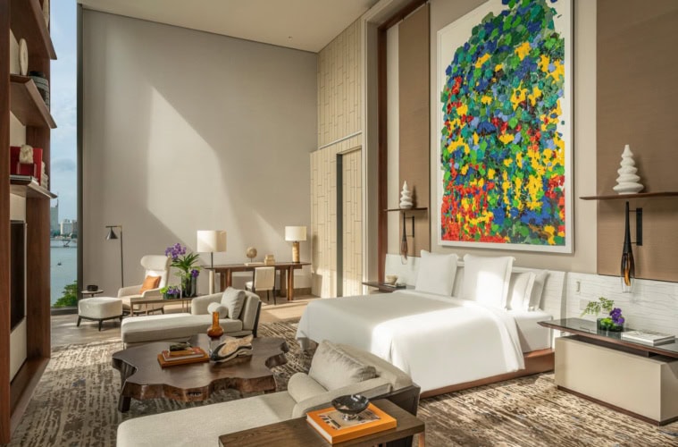 Blick in eine große, moderne Hotelsuite, mittig ein breites Bett, darüber ein gut zwei Meter hohes, modernes Gemälde. Vor dem Bett ein aus einem Baumstamm geschnittener Couchtisch, zu den Seiten je eine Recamiere.