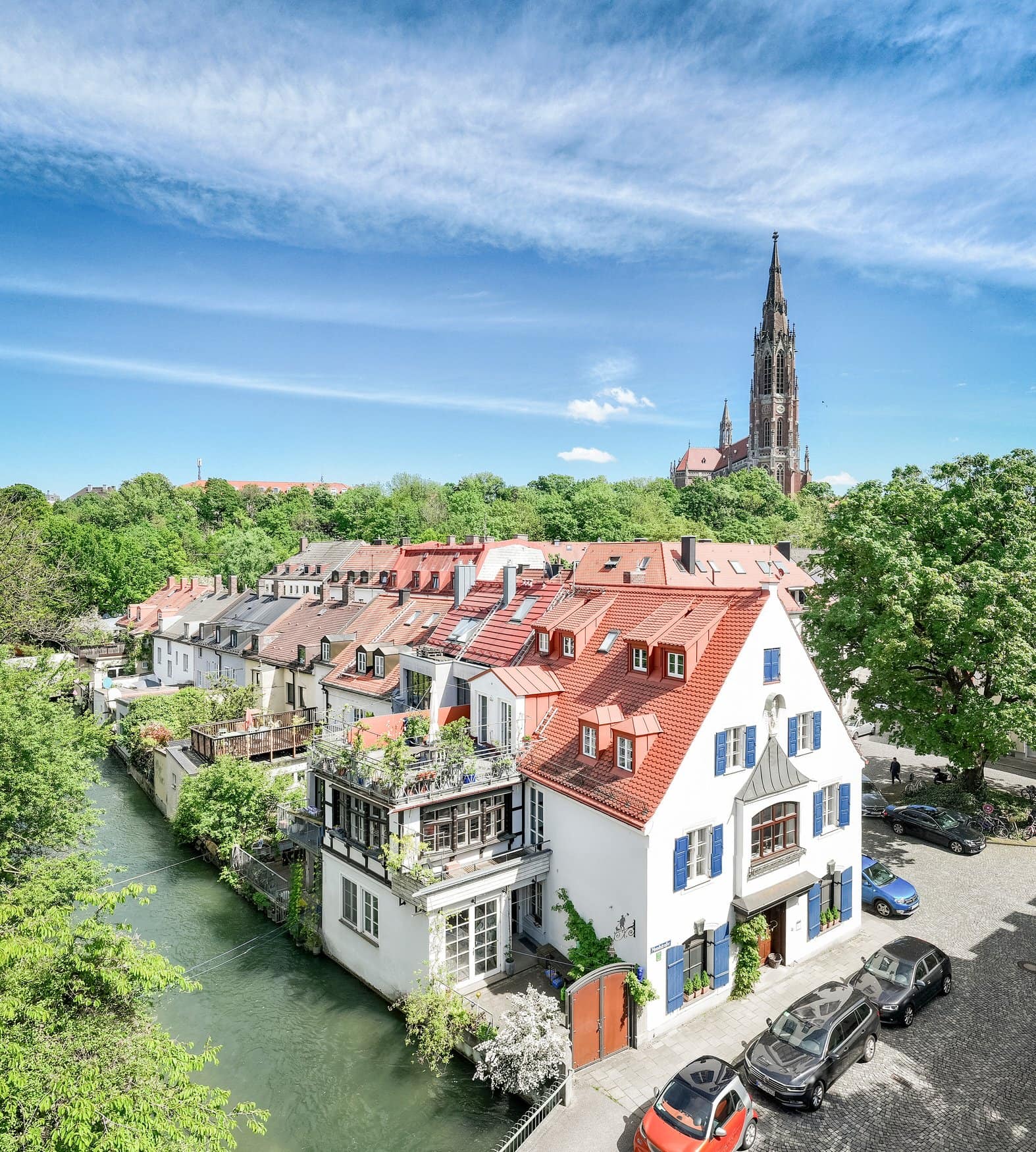 Blick von oben auf historische Häuser mit roten Dächern und blauen Fensterläden am Auer Mühlbach in Giesing, im Hintergrund der neugotische Turm der Mariahilfkirche.