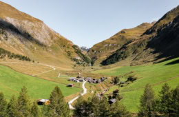 Die Fane Alm in Gitschberg Jochtal in Südtirol. Ein paar Häuser stehen in einem Tal neben einem kleinen Bach inmitten von Wiesen bedeckten Bergen.