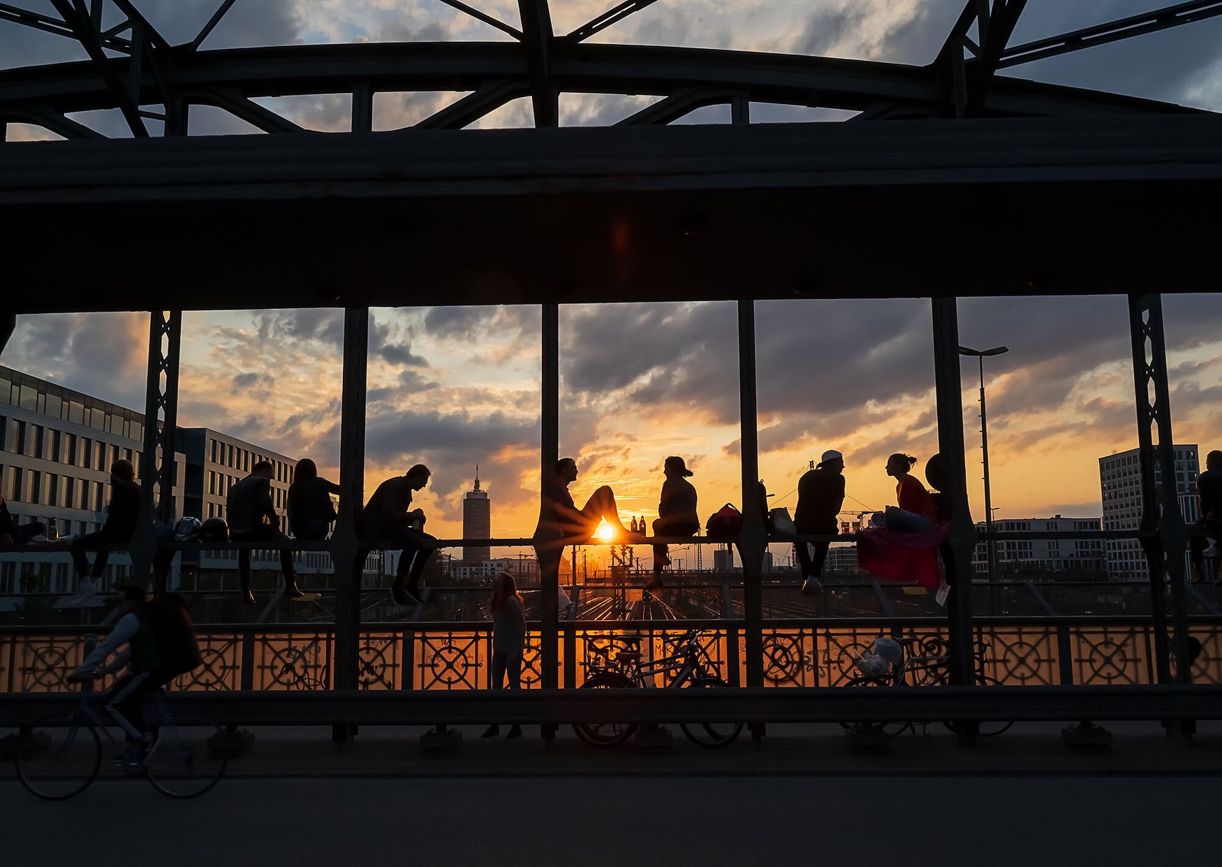 Silhouetten von Menschen, die auf dem Geländer der Hackerbrücke in München sitzen und den Sonnenuntergang beobachten, der den Himmel in Orange taucht; im Vordergrund Radfahrende und Fußgänger.