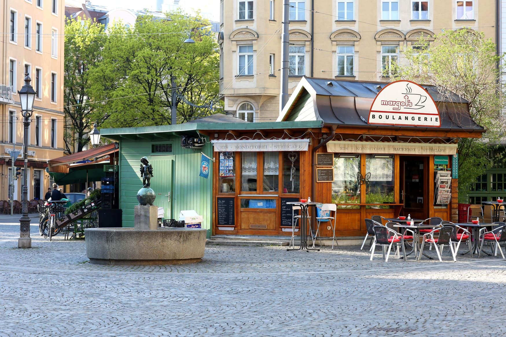 Kleiner Stand mit Brunnen und Bronzestatue auf dem Wiener Platz in Haidhausen, daneben die Holzfassade der Bäckerei und Konditorei Margot's Boulangerie mit Außentischen auf dem Kopfsteinpflaster.