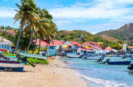 Bunte Fischerboote am Sandstrand von Bourg des Saintes, Îles des Saintes, mit pastellfarbenen Häusern und Palmen im Hintergrund