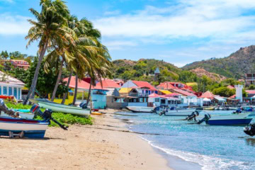 Bunte Fischerboote am Sandstrand von Bourg des Saintes, Îles des Saintes, mit pastellfarbenen Häusern und Palmen im Hintergrund