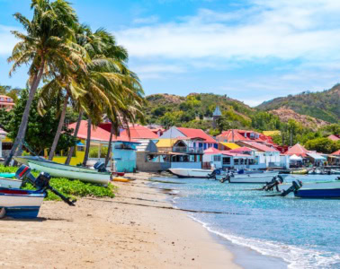 Bunte Fischerboote am Sandstrand von Bourg des Saintes, Îles des Saintes, mit pastellfarbenen Häusern und Palmen im Hintergrund