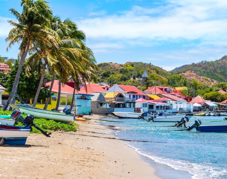 Bunte Fischerboote am Sandstrand von Bourg des Saintes, Îles des Saintes, mit pastellfarbenen Häusern und Palmen im Hintergrund