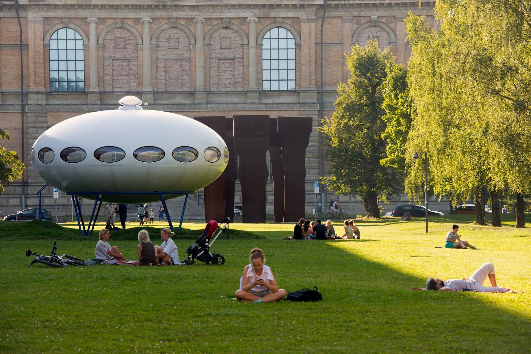 Das futuristische Futuro-Haus auf blauen Stelzen steht auf der Wiese vor der Alten Pinakothek in München; im Sonnenlicht sitzen und liegen Menschen auf dem Rasen.