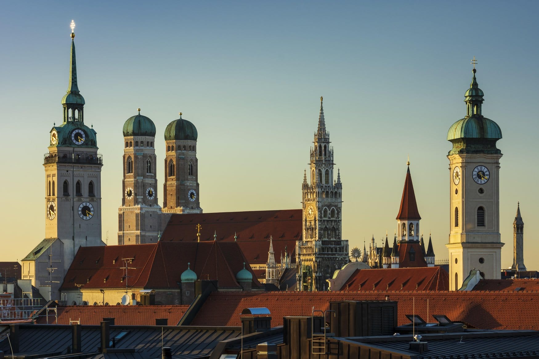 Stadtpanorama der Münchner Innenstadt im goldenen Abendlicht mit den Türmen von Peterskirche, Frauenkirche, Neuem Rathaus und Heilig-Geist-Kirche über den roten Dächern der Altstadt.