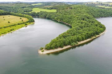 Vogelperspektive auf eine bewaldete Halbinsel im Obersauer-Stausee, umgeben von Wasser, Wäldern und Wiesen im Naturpark Obersauer in Luxemburg.