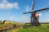 Ein gelb-blauer Zug der niederländischen Bahngesellschaft NS fährt an einer historischen Windmühle durch die niederländische Landschaft.