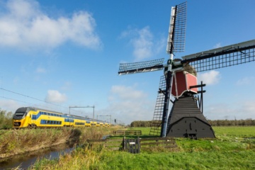 Ein gelb-blauer Zug der niederländischen Bahngesellschaft NS fährt an einer historischen Windmühle durch die niederländische Landschaft.