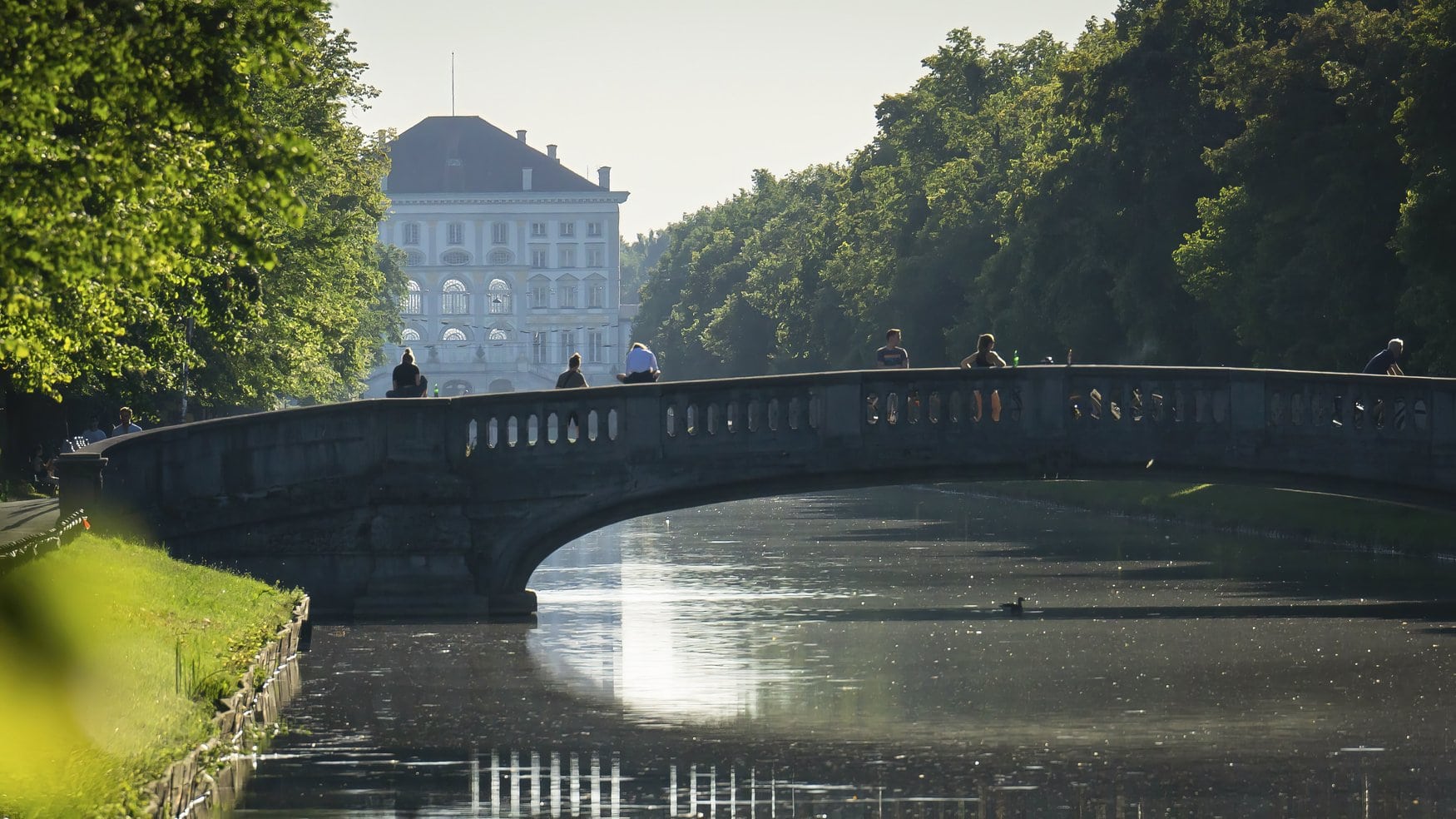 Brücke über den Nymphenburger Kanal in frühmorgendlichem Gegenlicht; auf der Balustrade sitzen und stehen Menschen, im Hintergrund zeichnet sich das Schloss Nymphenburg im Dunst ab.