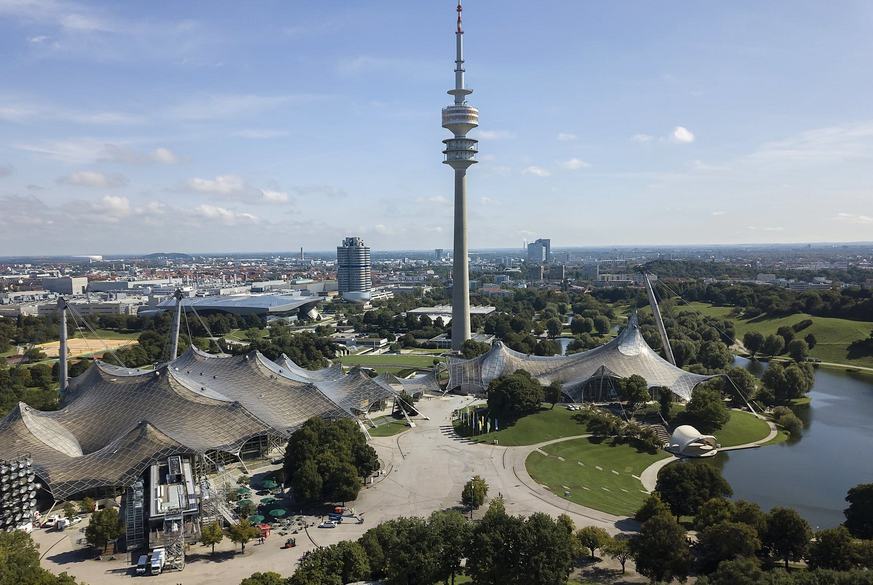 Luftaufnahme des Münchner Olympiageländes mit dem charakteristischen Zeltdach des Olympiastadions, dem Olympiaturm und dem Olympiasee sowie dem BMW-Vierzylinder-Hochhaus im Hintergrund.