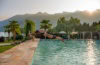 Ein Mann springt kopfüber vom Rand eines Hotelpools ins Wasser. Am Beckenrand sitzt eine Frau und schaut ihm zu. Im Hintergrund sind die Alpen sichtbar.