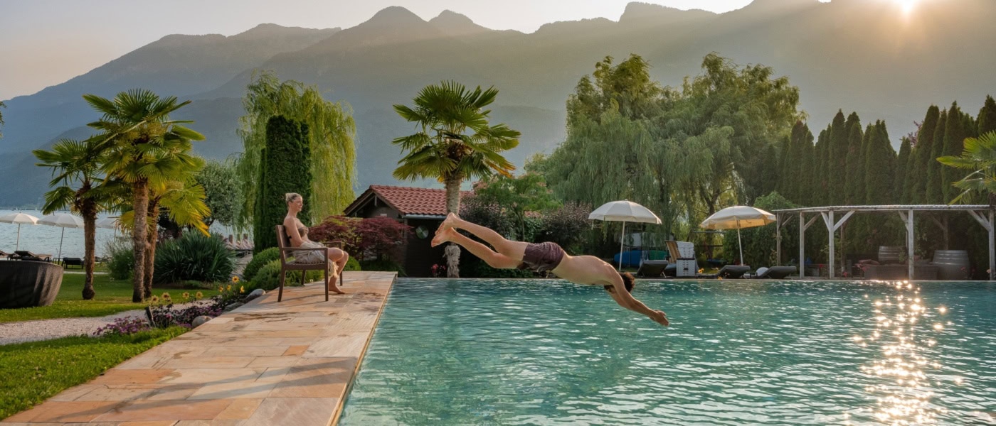 Ein Mann springt kopfüber vom Rand eines Hotelpools ins Wasser. Am Beckenrand sitzt eine Frau und schaut ihm zu. Im Hintergrund sind die Alpen sichtbar.