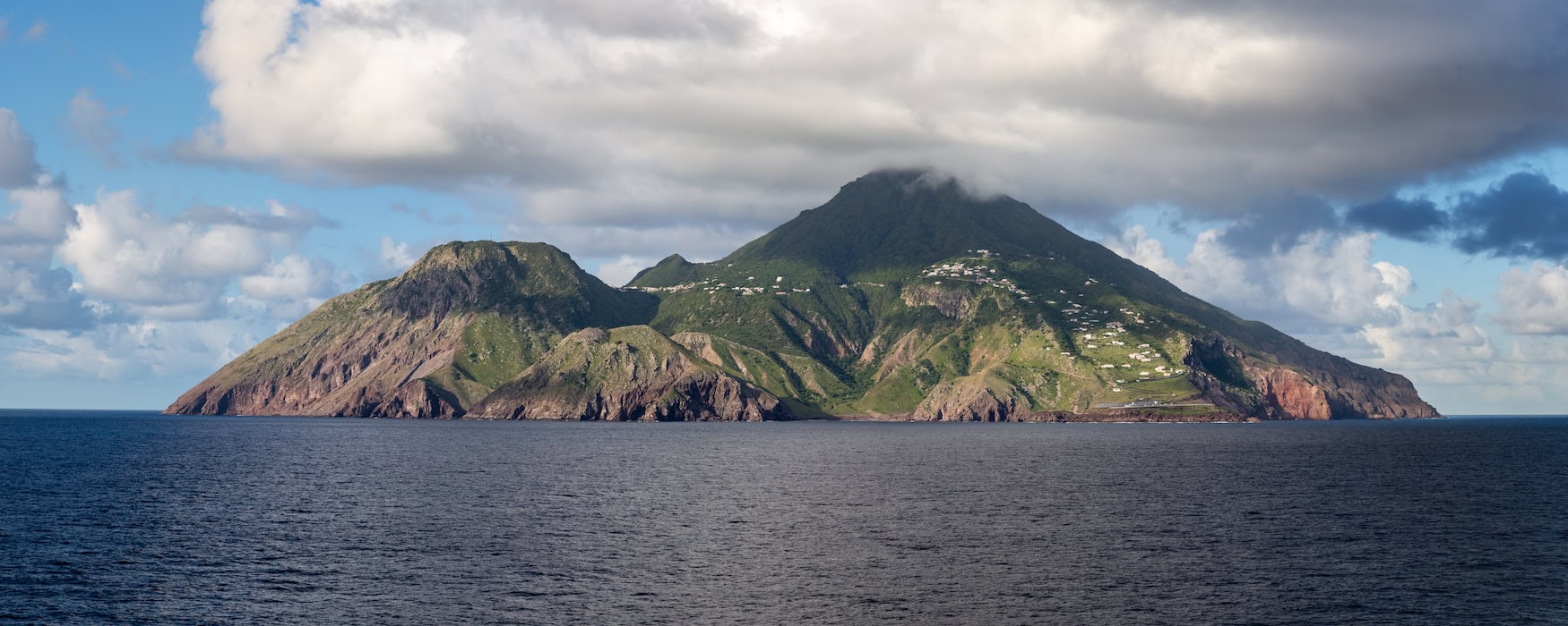  Panoramaaufnahme der Karibikinsel Saba vom Wasser aus: steile Felshänge, grüner Vulkankegel mit vereinzelten Häusern, tiefblauer Atlantik im Vordergrund