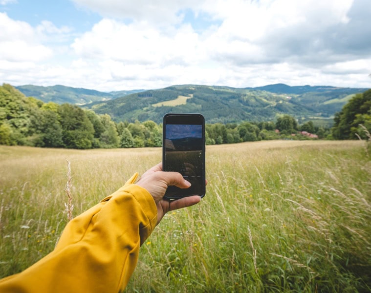 Eine Hügellandschaft mit gelben Feldern und grünen Wäldern. Eine Person hält ein Smartphone vor die Kamera und nimmt ein Foto auf.