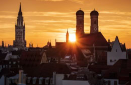 Dramatischer Sonnenuntergang über der Münchner Innenstadt mit den Silhouetten der Frauenkirche mit ihren Zwiebeltürmen und dem Turm des Neuen Rathauses vor leuchtendem Orangehimmel.