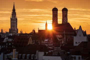 Dramatischer Sonnenuntergang über der Münchner Innenstadt mit den Silhouetten der Frauenkirche mit ihren Zwiebeltürmen und dem Turm des Neuen Rathauses vor leuchtendem Orangehimmel.