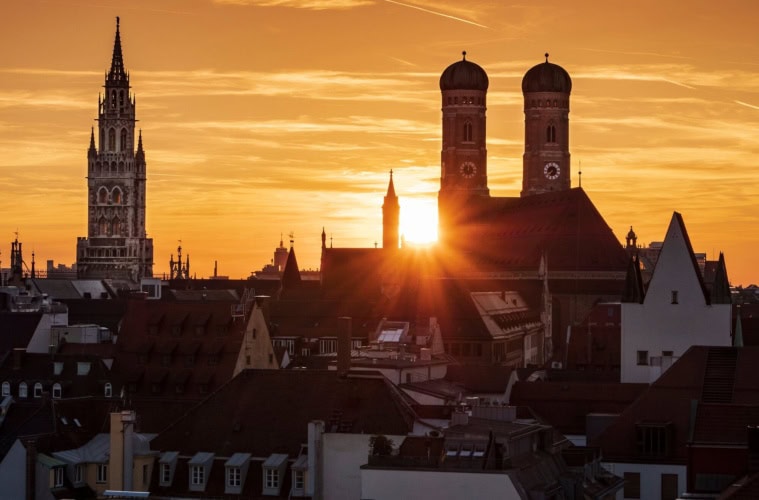 Dramatischer Sonnenuntergang über der Münchner Innenstadt mit den Silhouetten der Frauenkirche mit ihren Zwiebeltürmen und dem Turm des Neuen Rathauses vor leuchtendem Orangehimmel.