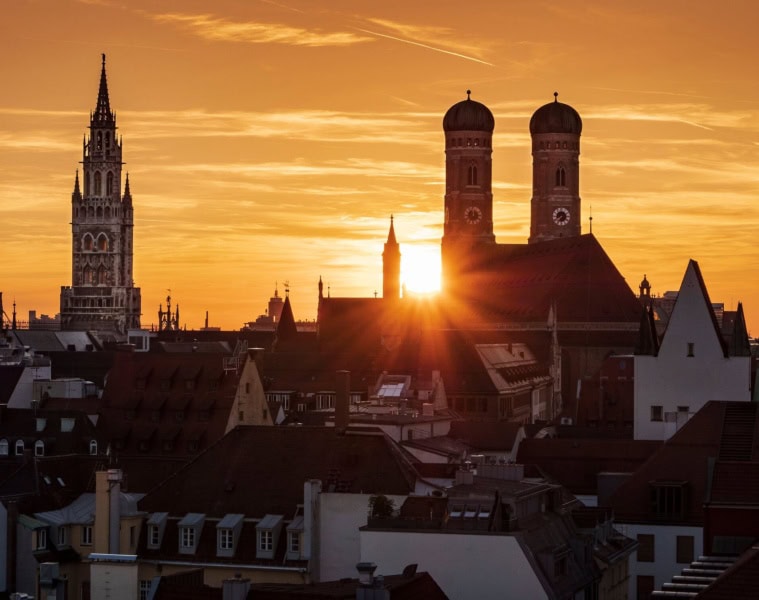 Dramatischer Sonnenuntergang über der Münchner Innenstadt mit den Silhouetten der Frauenkirche mit ihren Zwiebeltürmen und dem Turm des Neuen Rathauses vor leuchtendem Orangehimmel.