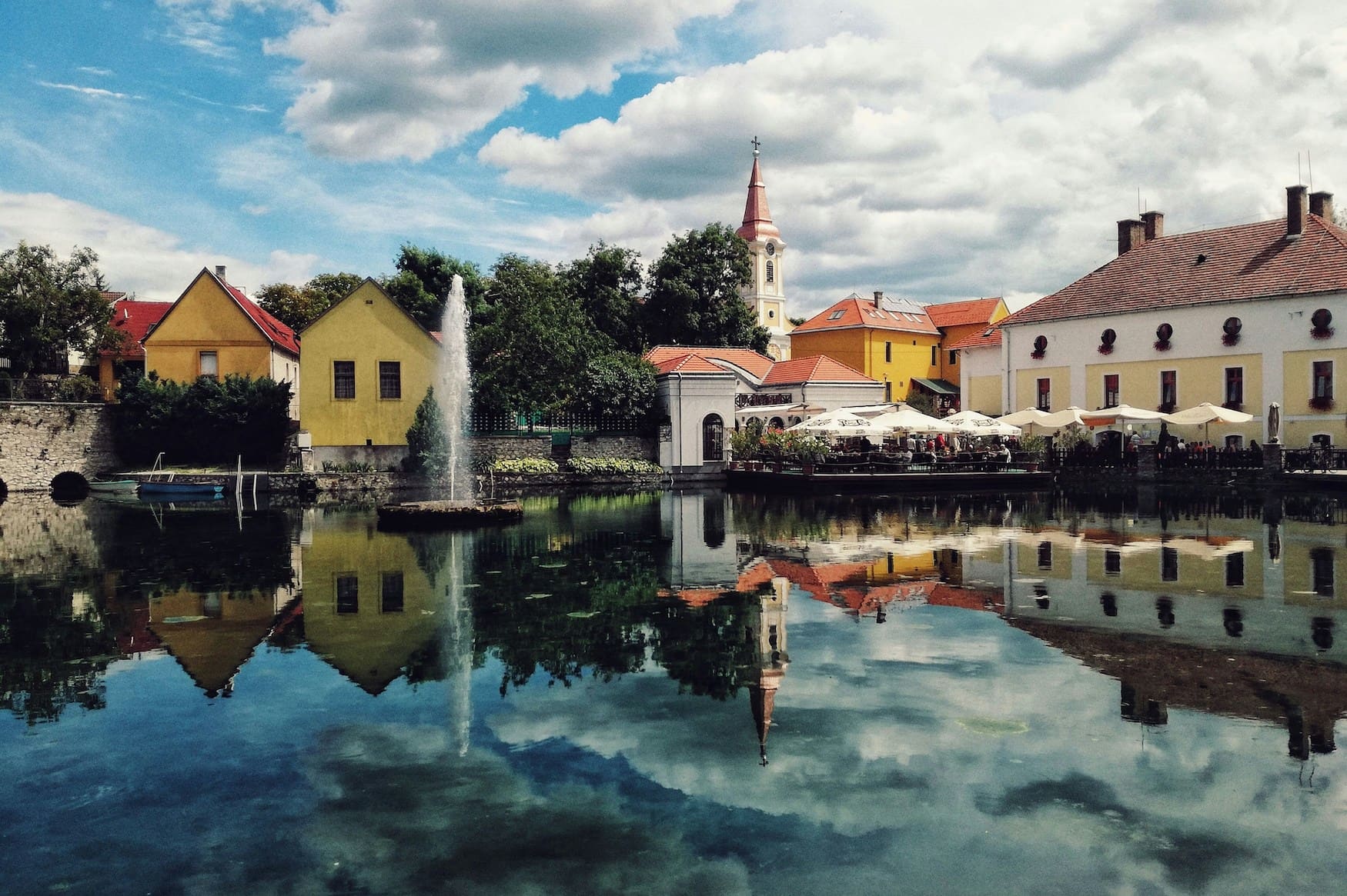Blick über den Stadtteich von Tapolca in Ungarn mit Springbrunnen, bunten Häusern und der Stadtkirche – Spiegelung im ruhigen Wasser