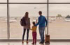 Ein Mann, eine Frau und ein Kind im Grundschulalter stehen mit ihrem Handgepäck an einer großen Scheibe enes Flughafens und betrachten die abhebenden Flugzeuge.