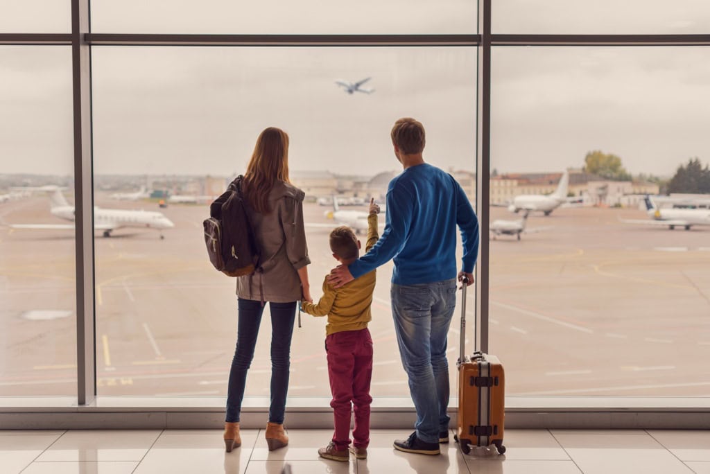 Ein Mann, eine Frau und ein Kind im Grundschulalter stehen mit ihrem Handgep&auml;ck an einer gro&szlig;en Scheibe enes Flughafens und betrachten die abhebenden Flugzeuge.